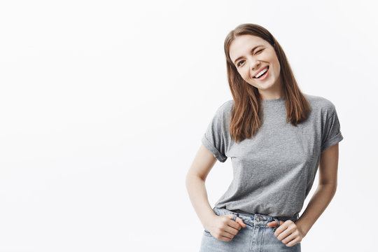 Close up of handsome confident brunette european student girl in casual grey t-shirt and jeans winking, looking in camera with cheerful face expression, smiling brightfully.