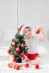 Boy in red Santa hat decorates a small Christmas tree balls