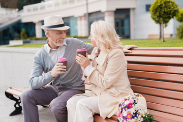 Obraz premium Two pensioners are sitting on a bench with a glass of coffee in their hands. They look at each other with tenderness
