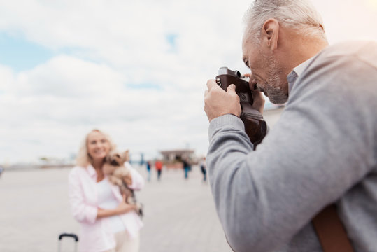 An elderly couple is walking. A woman poses with a dog in her arms, a man takes pictures of her on a film camera