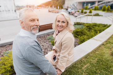 An elderly couple is sitting on the edge of a flower bed. They smile at each other