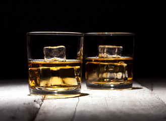 Two glasses of whiskey with ice cubes served on white wooden planks.