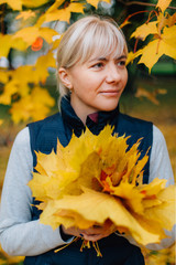 Pretty young blonde woman in waistcoat  holding yellow maple leaves in colorful autumn park.