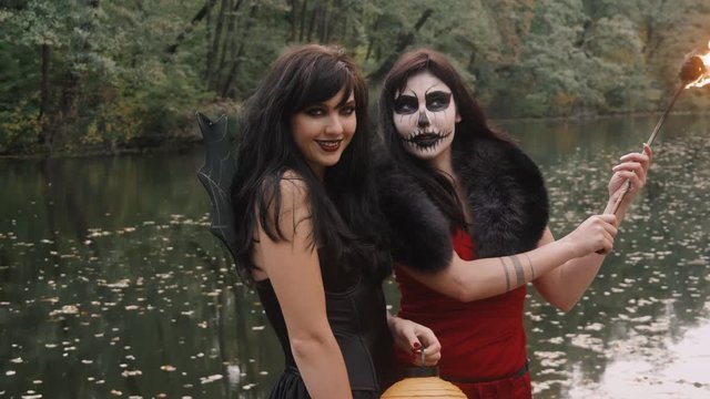 Young halloween girl friends in black and red witch dress having fun in the forest park with traditional autumn holiday symbol of orange spooky pumpkin outdoor.