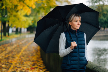 Pretty young blonde sad woman with umbrella in waistcoat in colorful autumn park. Depressed mood.