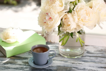 Cup of tea and vase with beautiful peony flowers on windowsill
