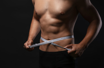 Sporty young man measuring his waist on dark background
