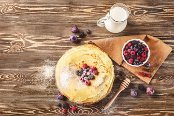 Stack of delicious thin pancakes served with berries and sugar powder on plate against wooden background