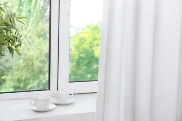 Window with beautiful white curtains and two cups on sill