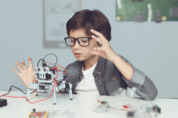 A small nerd in glasses is holding a robot. He enthusiastically looks at his creation