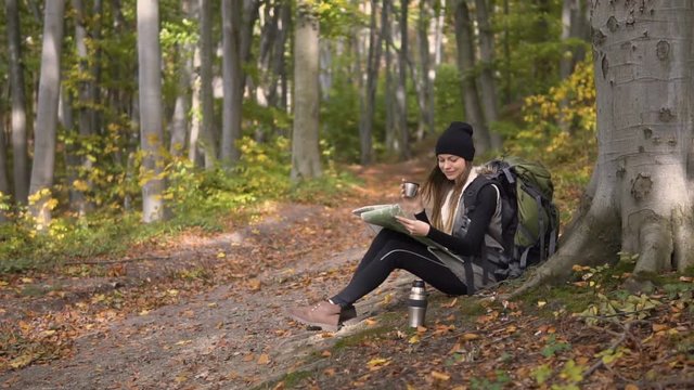 Smiling long hair girl, with green backpack, looking at the map and having tea while resting in the forest