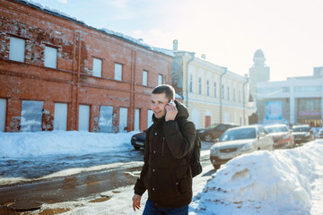 Young man with a backpack goes on the winter city and speaks by phone.