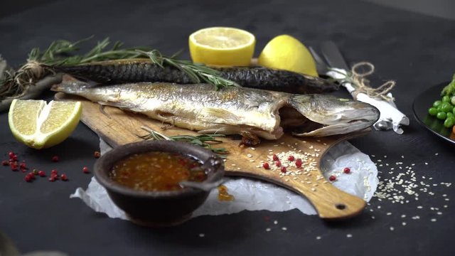 Fried fish on a cutting board, next to a rosemary, lemon, side sausage and a plate with vegetable garnish