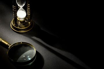 Old vintage hourglass with magnifying glass with shadow on dark background with free space