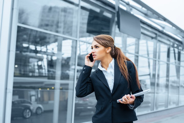 Successful businesswoman or entrepreneur using a digital tablet computer and talking on cellphone standing in front of his office.