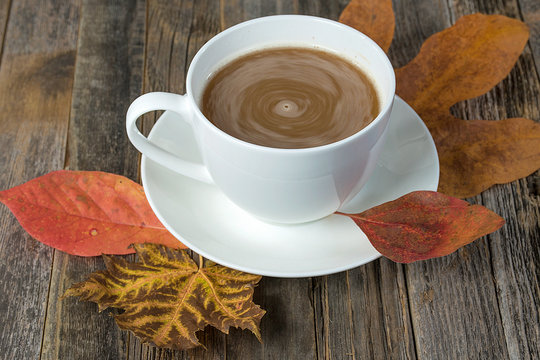 swirling cream and coffee in white cup on autumn leaves and rustic wood