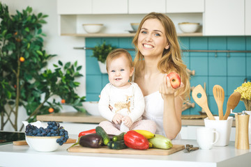 Obraz na płótnie Canvas Young mother looking at camera and smiling, cooking and playing with her baby daughter in a modern kitchen setting. Healthy food concept.