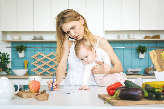 Young Mother With Her Baby Daughter In A Modern Kitchen Setting. Young Attractive Cook Woman Desperate In Stress, Tired.