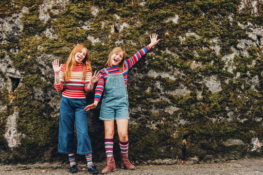 Outdoor portrait of two funny preteen girls wearing denim shorts, stripe roll neck pullovers and long socks, posing against moss wall, fashion for teens