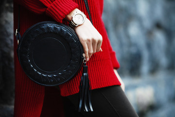 Fashionable woman posing in street, near wall, holding black round bag, wearing wrist watch,...