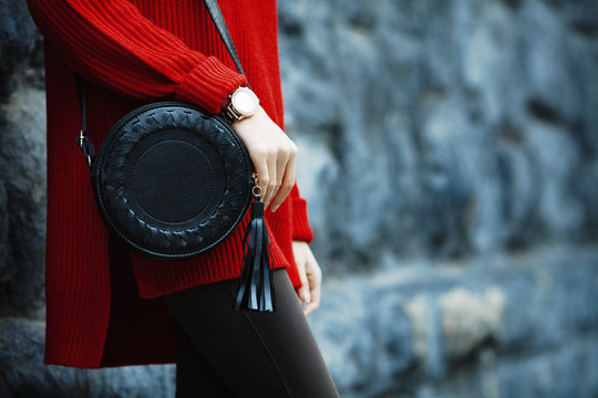 Fashionable woman posing in street, near wall, holding black round bag, wearing wrist watch, stylish red oversized sweater. Female fashion concept. Copy, empty space for text.