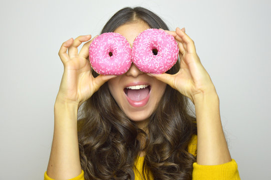 Close-up portrait of long-haired smiling girl having fun with sweets isolated on gray background. Attractive young woman with long hair posing with pink donuts in her hands
