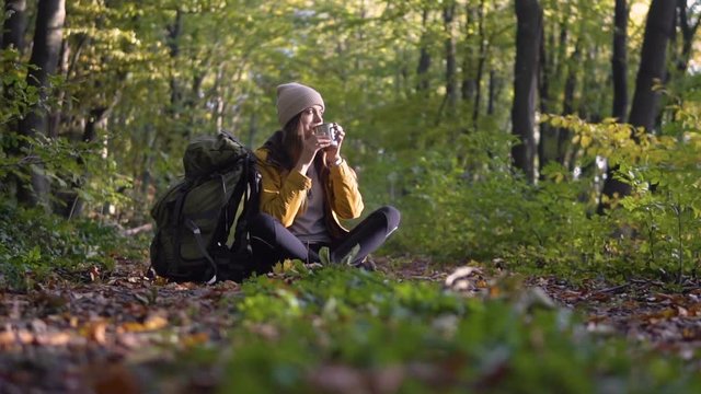 Female skinny tourist is having tea and taking the map out of the backpack in the wood, slowmotion on fall day