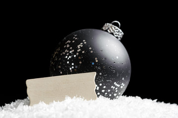 Blank wooden sign and black and silver glittering ornament sitting in snow