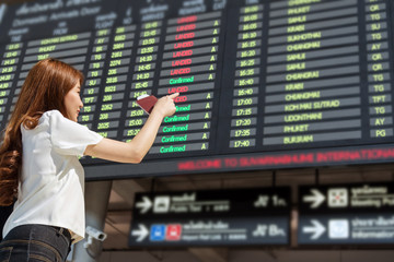 Asian woman traveler holding the passport and pointing at the flight information screen in modern an airport, lifestyle travel and transportation concept.