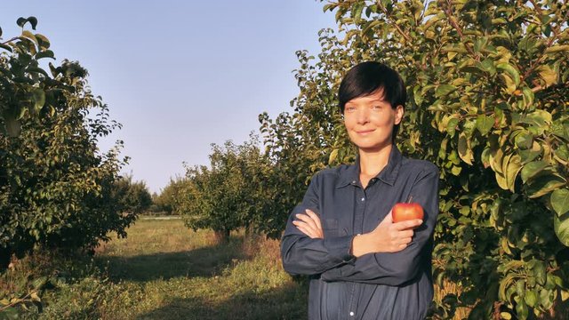 Adult woman posing in garden with trees holding red apple. Happy cheerful fruit grower wearing in blue working overalls in summer season sunny day