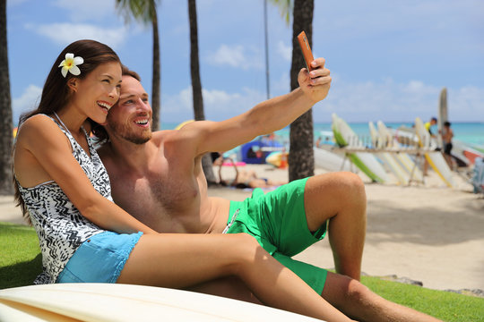 Selfie couple taking phone picture on Hawaii Waikiki beach travel vacation. Young people surfers lifestyle relaxing in Honolulu city after surf session with surfboards. Asian girl, Caucasian man.