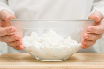 Chef's hands holding a glass bowl of cottage cheese on the wooden board for pie, cake, sweet dessert or pancakes in the kitchen. Preparation for cooking. Food concept.
