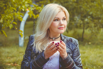 Middle age stylish blonde hair pretty woman dressed in leather black jacket on a cozy picnic in autumn garden with philosophical mood. Lady with pretty accessorize and casual style on a nature