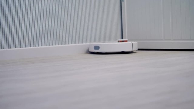 A round cleaning robot slowly moves along a bedroom wall. 