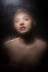 Portrait of a bald girl behind a wet glass on a black background.