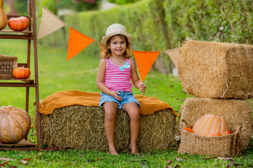 Naklejka na ściany i meble Cute little girl sitting among hay bales and pumpkins in the garden