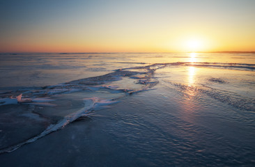 Winter landscape with cracks on the frozen lake near the shore at sunset