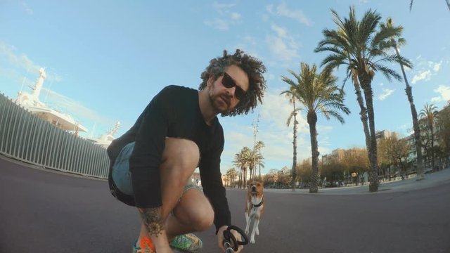 Cool curly man rides his longboard next to sea port at early morning with happy basenji dog running nearby.He spins around, holds selfie stick to film from low angle, covers camera with hand in end