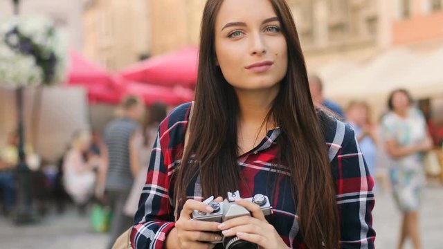 Portrait of attractive caucasian woman standing in the middle of the street with vintage camera and smiling at the camera.