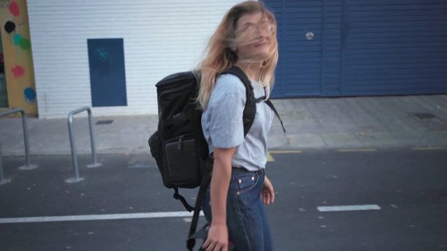 Pretty smiling blonde tourist girl walking in gentrificated district of town with big black backpack and professional camera and takes photos on her way at evening time.