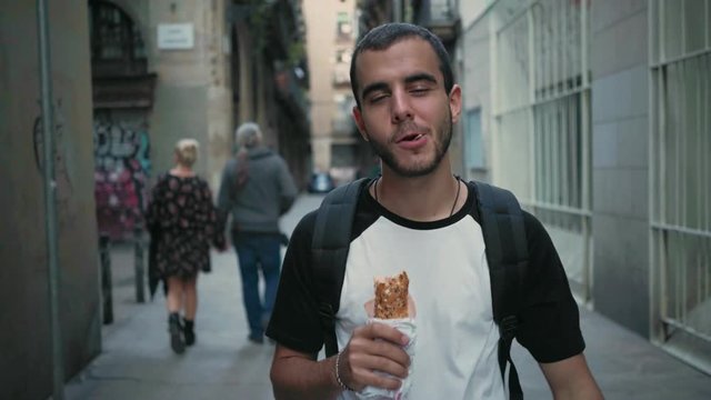 Young handsome man explores city attractions, eats take away sandwich food on the go, chews and bites huge sub or BLT, fulfills his appetite, busy to stop at slowfood restaurant