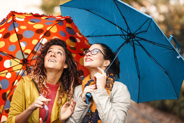 Two female best friend walks at the city street with umbrellas above head.They smiling and making...