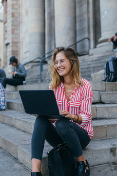 Happy cheerful woman in retro vintage prescription glasses works on laptop while sitting on porch or stairs of university or museum, modern business lady or student works from home