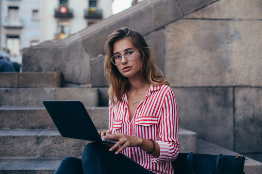 Serious and concentrated young woman, in fashion trendy outfit sits on stairs of office building or university, works, chats or types on her laptop, new project, looks into camera