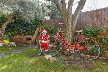 Fototapeta na wymiar Kid dressed as Santa Claus sit in a swing in the garden. Outdoor. Christmas time.