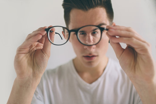 Closeup portrait of young man with glasses. He has eyesight problems and is squinting his eyes a little bit. Handsome guy is holding his eyeglasses right in front of camera with one hand. The concept