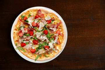 Prosciutto pizza with cherry tomatoes and rucola on dark brown table from above.