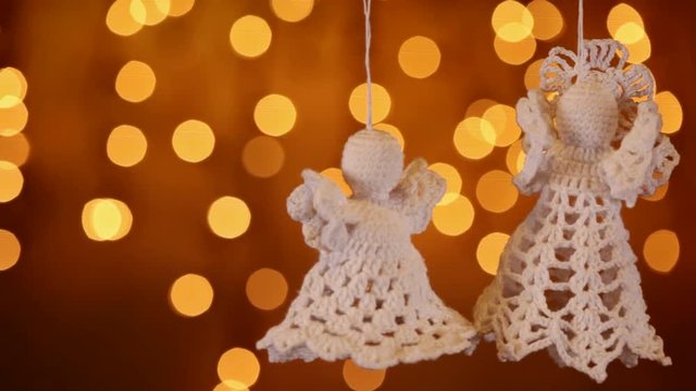 Two crocheted xmas angel decorations hanging in front of golden blurry christmas lights - camera slide