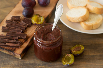 Plum chocolate jam in a glass jar, fresh plums and pieces of chocolate on a wooden surface.