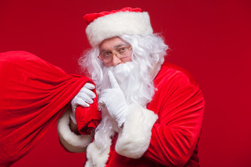 Portrait of Santa Claus with huge red sack keeping forefinger by his mouth and looking at camera